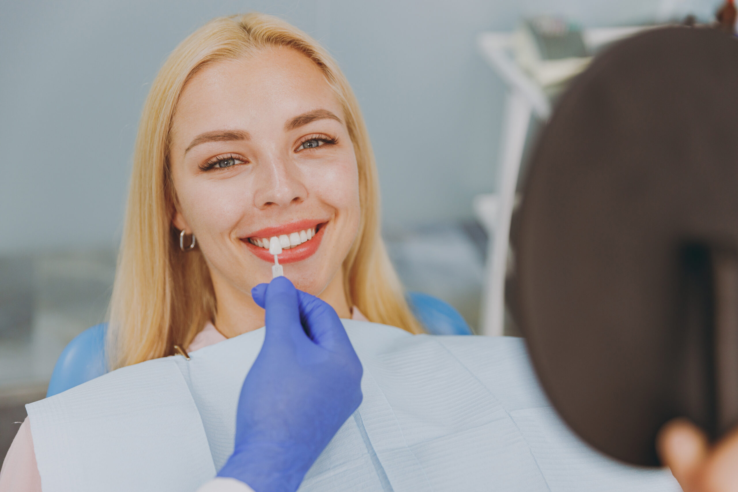 Young smiling calm happy woman 20s choose veneers enamel color compare teeth shade with stomatologist look at result mirror sit at dentist office chair indoor modern cabinet Enamel whitening treatment