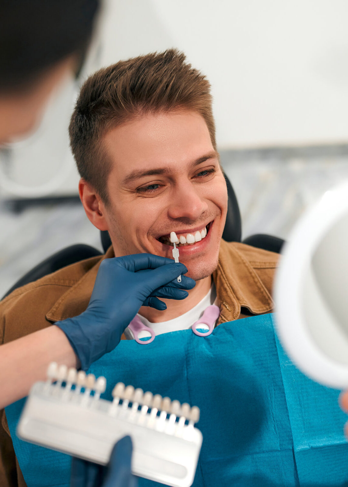 Medicine, dentistry and healthcare concept - closeup of a dentist with tooth color samples choosing a shade for a male patient's teeth in a dental clinic looking at a mirror.
