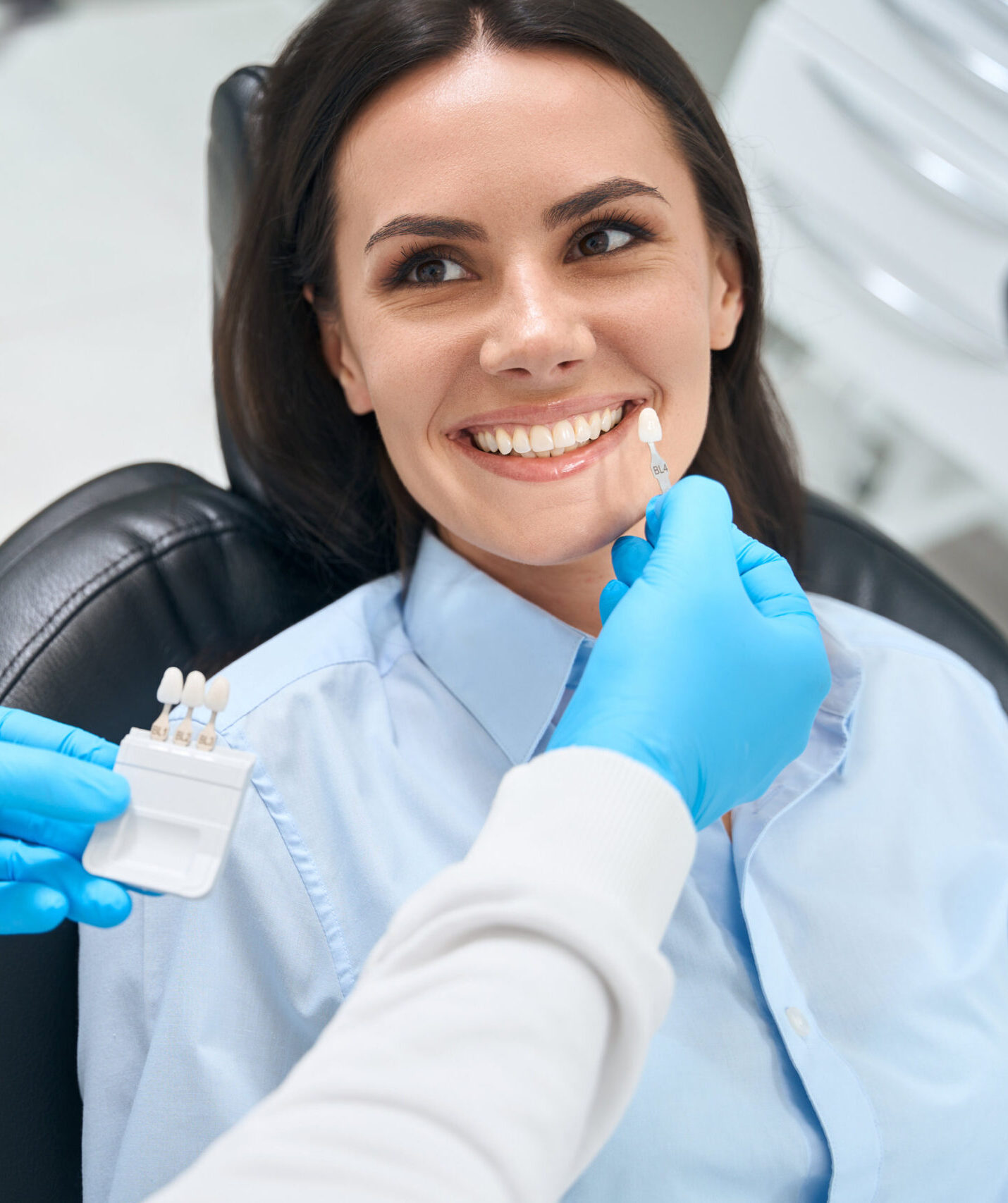 High-qualified dental technician choosing proper size and color of dental crown using some template, smiling woman client sitting in cozy orthodontic chair