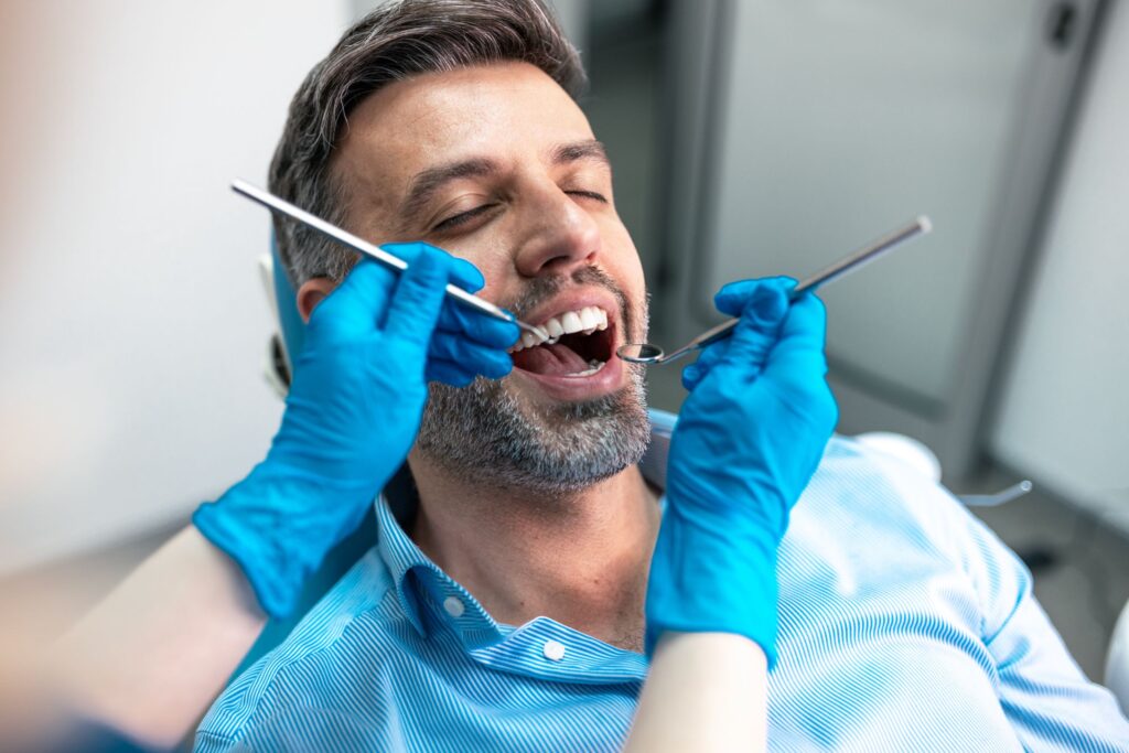 Dentist using dental instruments to examine a smiling male patient's open mouth