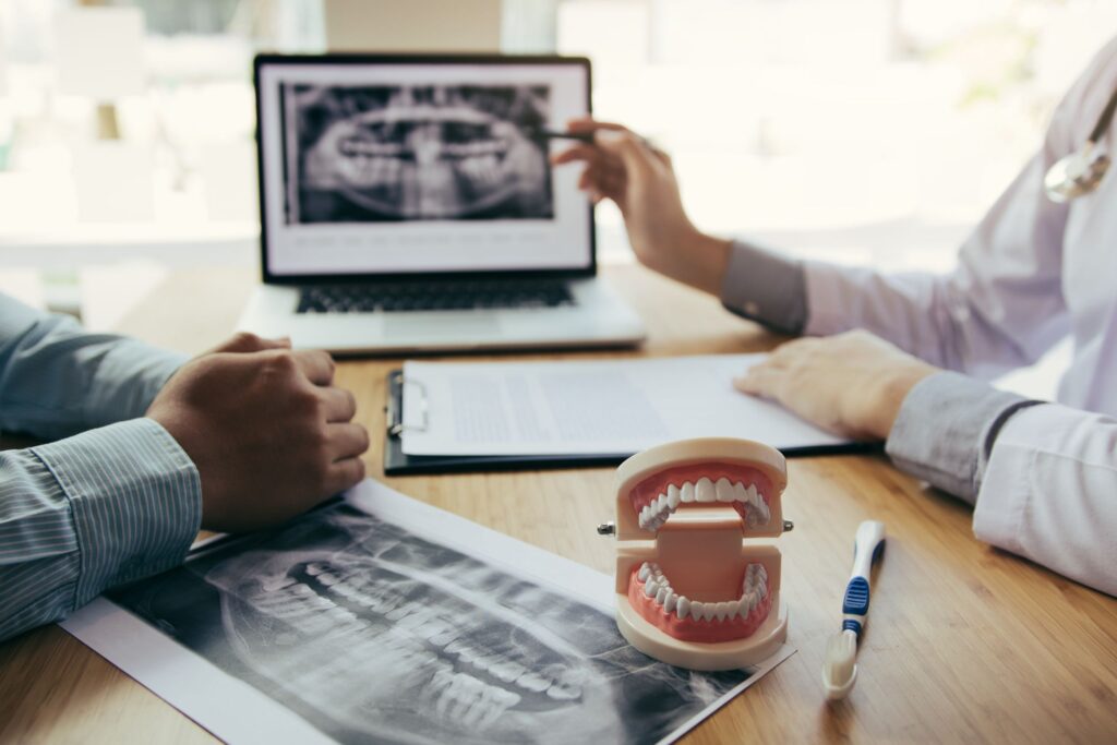 Dentist pointing to a panoramic dental X-ray on a laptop next to a tooth model and physical X-ray film